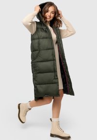Grüne Daunenweste mit Kapuze, mit gestepptem Design, seitlichen Taschen und pinken Akzenten. Getragen über einem cremefarbenen Pullover, kombiniert mit beigen Schnürstiefeln.