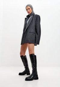 Botas negras hasta la rodilla con suela gruesa, combinadas con un blazer gris de gran tamaño y un suéter de cuello alto, sobre un fondo blanco liso.