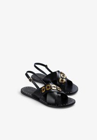 Sandali neri in pelle con cinturini incrociati, dettagli circolari dorati e una suola piatta caratterizzata da piccole perforazioni. Design slingback per una vestibilità sicura.
