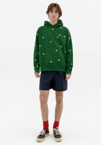 Green hoodie with white floral patterns, front pocket, and drawstring hood. Paired with navy shorts and red socks, featuring dark sneakers.