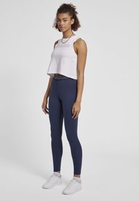 Top corto bianco con logo stampato "HUMMEL", abbinato a leggings blu navy con fascia in coordinato. La modella indossa sneakers bianche.
