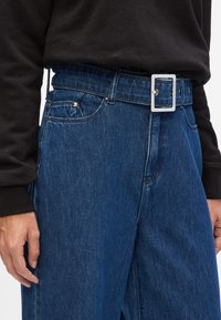 Jean en denim bleu foncé avec une coupe décontractée, comprenant une large ceinture, une boucle en métal et un subtil détail de poche. Texture lisse, design classique.