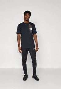 Camiseta negra de cuello redondo con gráfico "JUST DO IT" en el lado izquierdo y logo azul; combinada con pantalones deportivos negros y zapatillas negras.