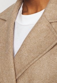Abrigo de mezcla de lana beige con un acabado texturizado, con un cuello con muescas y líneas limpias, superpuesto a una camiseta blanca de cuello redondo.