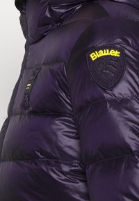 Paarse pufferjas met glanzende textuur. Voorzien van een geel logo-embleem en een ritssluiting aan de zijkant. Warme, geïsoleerde uitvoering.