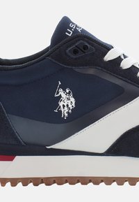 Zapatillas de deporte azul marino con acentos blancos y suela de goma texturizada. Presenta un logo de tres jinetes en el lateral y materiales contrastantes.