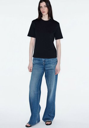 Kobieta stojąca, ubrana w prostą czarną koszulkę z krótkim rękawem, szerokie niebieskie jeansy i czarne sandały z odkrytymi palcami, na białym tle.