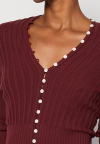 Tummanpunainen ribbineulottu paita V-pääntiellä, jossa koristeelliset valkoiset napit edessä ja aaltoileva reunus pääntiellä.