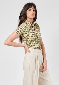 Camicia a colletto con maniche corte, caratterizzata da un motivo verde con dettagli viola e bianchi. Pantaloni beige a vita alta con pieghe frontali.