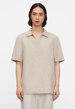 Persona con capelli neri fino alle spalle, che indossa una camicia beige a maniche corte con colletto e pantaloni abbinati su uno sfondo semplice.