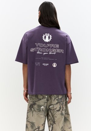Lilla oversized t-shirt med en hvid grafisk tryk på bagsiden, der viser teksten "DU ER STÆRKERE end du tror" og indviklede designs.