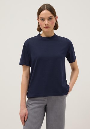 Vrouw met kort haar, draagt gouden creolen, een marineblauw kortermig shirt en grijze broek met krijtstreep, poseert met één hand in haar zak.