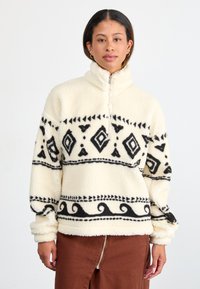 Fleece pullover in crème met zwart geometrisch patroon en golven. Heeft een halflange ritssluiting en omslagmouwen voor extra warmte.