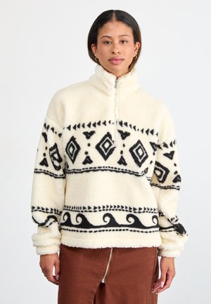 Pull en polaire crème avec un motif géométrique noir et des vagues. Il dispose d'un col zippé demi-long et de manches à revers pour plus de chaleur.