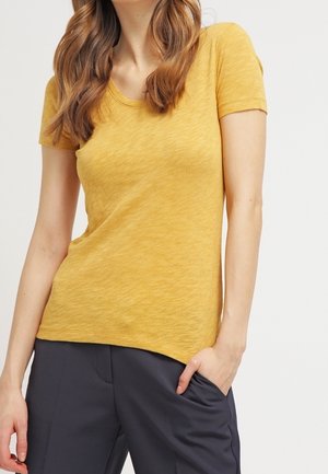 Kvinde iført en kortærmet sennepsgul t-shirt og mørkegrå bukser med den ene hånd i lommen foran en ensfarvet baggrund.