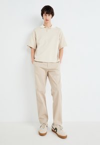 Beige poloing, kontrasztos gallérral, bézs chino nadrággal és világos edzőcipővel, sötétzöld kiegészítéssel társítva. Simas kifejezés, laza fit.