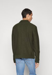 Veste en laine verte foncée avec un col, des coutures visibles et des manches longues, assortie à un jean bleu clair. Vue de dos affichée.