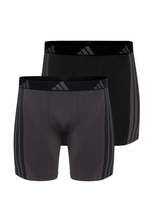 Muscular man standing barefoot wearing black athletic compression shorts with side stripes and logo on waistband against plain background.