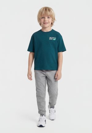 Niño rubio con camiseta verde azulado de New Balance, pantalones deportivos grises y zapatillas blancas, de pie y sonriendo contra un fondo liso.