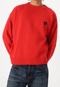 Pull rouge en maille avec un col rond, des poignets et un ourlet côtelés, comportant un petit motif de cœur noir sur le côté gauche de la poitrine.