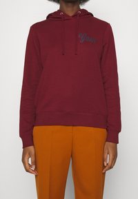 Sudadera en color burdeos con cordones. Cuenta con un logo bordado de "Gant". Tela suave, puños y dobladillo acanalados, combinada con pantalones naranjas.