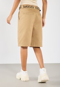 Shorts khaki en tissu texturé, présentant une ceinture à motif léopard, des poches arrière et une coupe décontractée. Portés avec des baskets blanches à semelles épaisses.