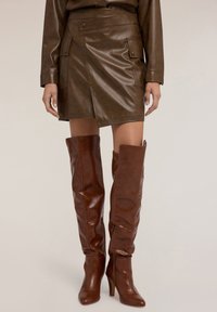 Gonna avvolgente in pelle marrone con tasche anteriori, abbinata a stivali marroni alti fino alla coscia con silhouette aderente e tacco a stiletto.
