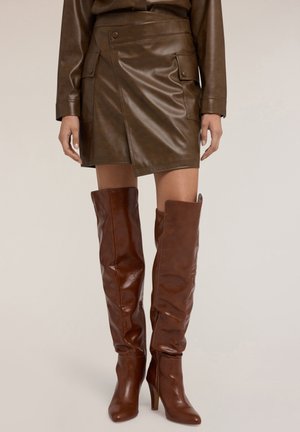 Gonna avvolgente in pelle marrone con tasche anteriori, abbinata a stivali marroni alti fino alla coscia con silhouette aderente e tacco a stiletto.