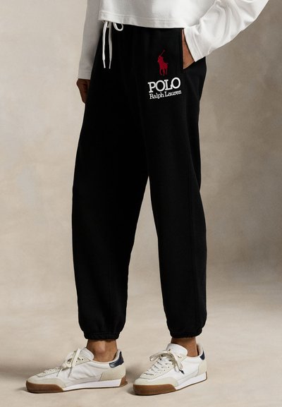 Sorte joggingbukser lavet af bomuld med elastisk taljebånd, med en rød logo og hvid tekst. Båret sammen med hvide og beige sneakers.