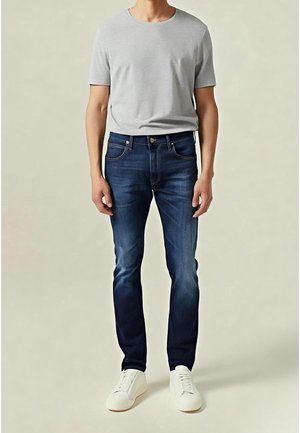 Man draagt nauwsluitende donkerblauwe spijkerbroek, lichtgrijs T-shirt met korte mouwen en witte sneakers, staand tegen een effen achtergrond.