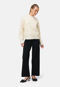 Kremfarvet sweater med teksturerede blomsterapplikationer, ribstrikkede ærmekanter og kant. Kombineret med brede, sorte bukser og sorte slingback-sko med sløjfer.