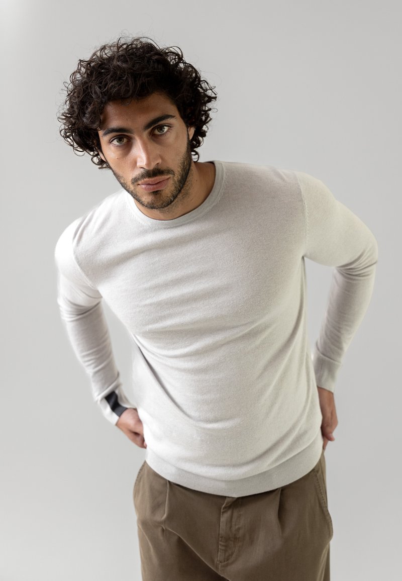 Hellbeige, langärmliger Pullover aus weichem Material mit taillierter Passform, konstruiert mit gerippten Bündchen und einem runden Ausschnitt. Getragen über braunen Hosen.