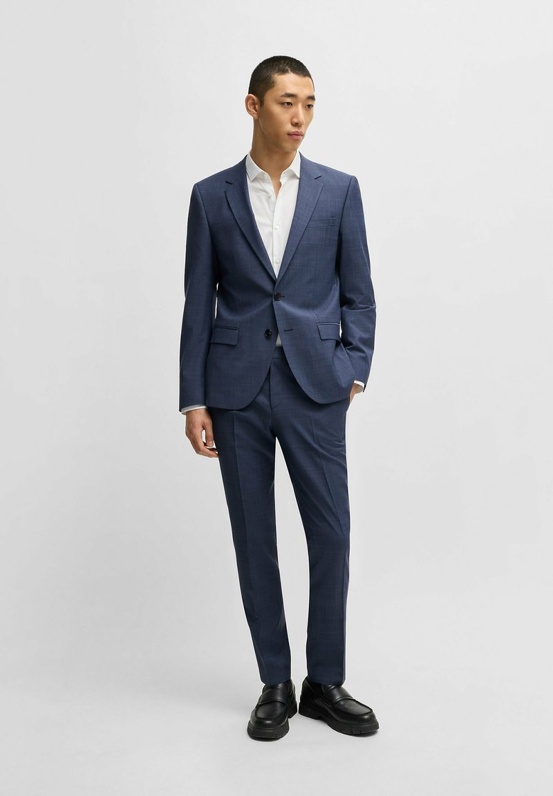Dunkelblauer Anzug mit Karomuster, einreihiger Blazer und schmal zulaufende Hose. Kombiniert mit einem weißen Hemd und schwarzen Schuhen.