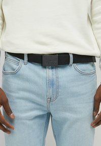 Sort stofbælte med en mat sort metalspænde prydet med det præget "Lee" logo. Båret med lysblå denim jeans og en hvid sweatshirt.
