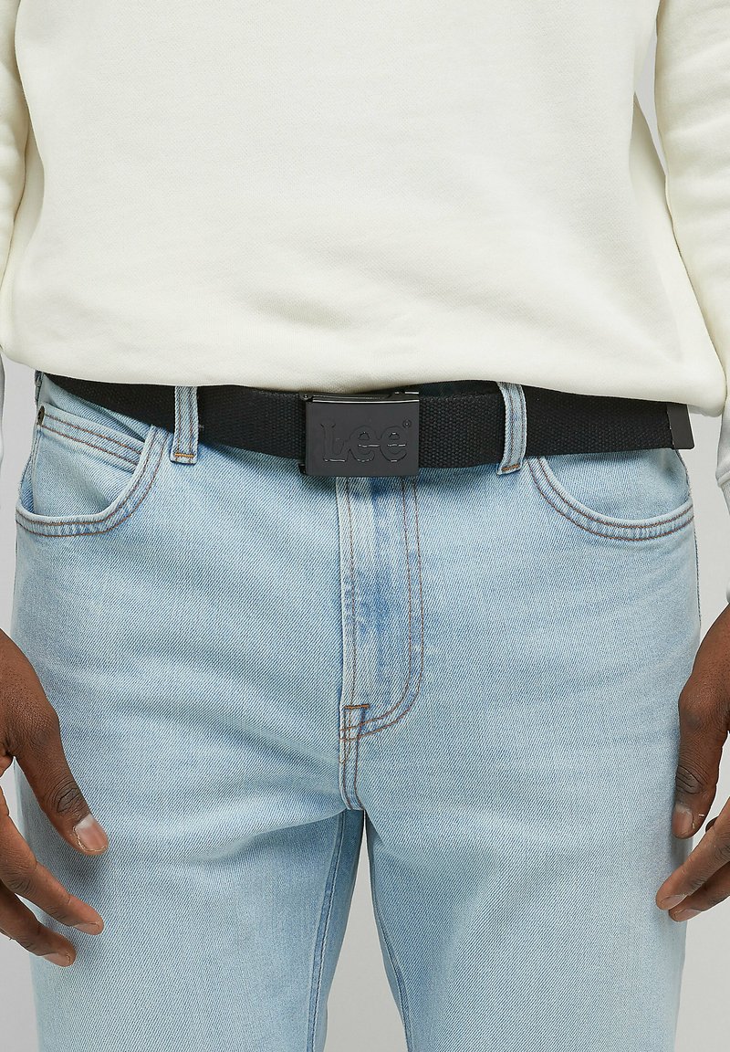 Sort stofbælte med en mat sort metalspænde prydet med det præget "Lee" logo. Båret med lysblå denim jeans og en hvid sweatshirt.