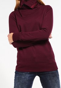 Sudadera burdeos con cuello alto y un bolsillo frontal cuadrado, hecha de tejido suave. Presenta mangas largas y una cintura ajustada.