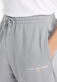 Vaaleanharmaat verkkarit, joissa on joustava vyötärö, nyörikiinnitys, sivutaskut ja valkoinen sekä kultainen "New Balance" -logo. Pehmeä kangas.