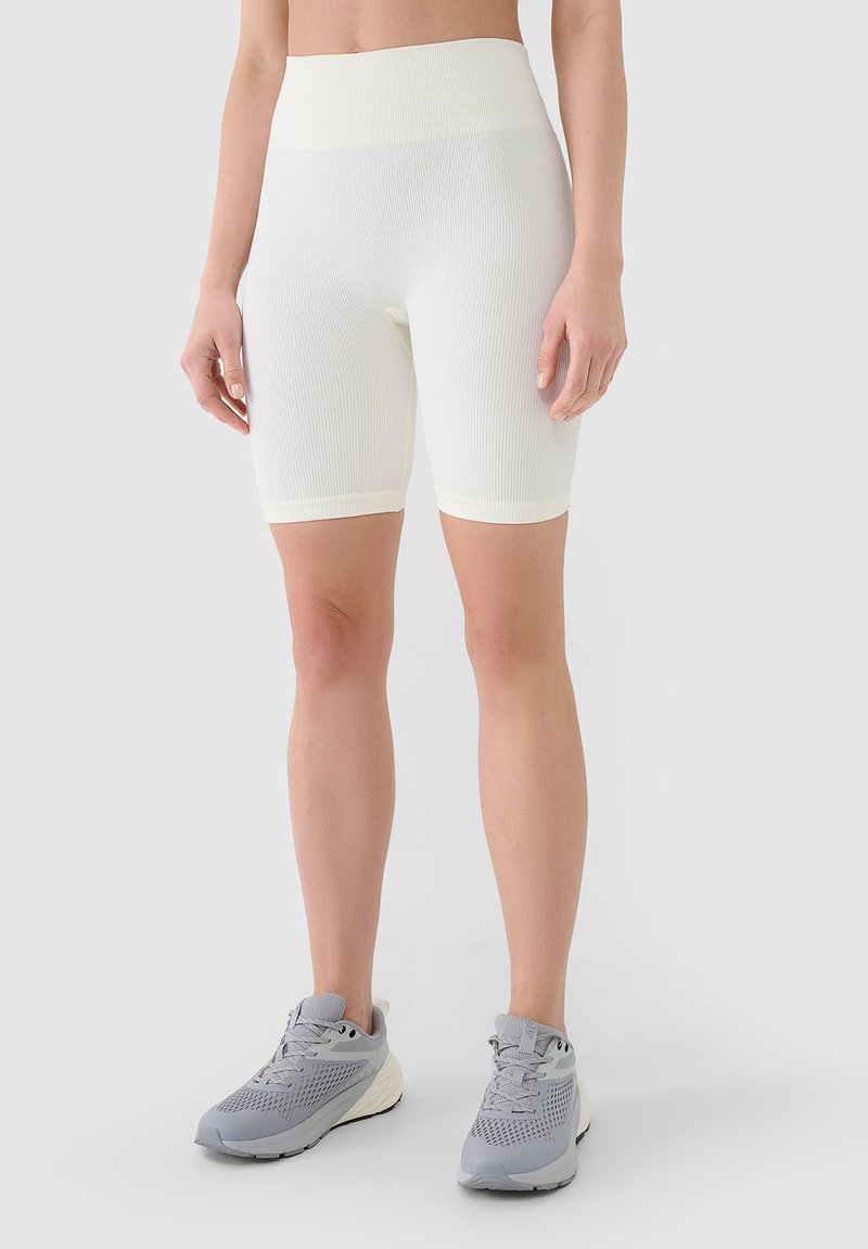Femme portant un cuissard blanc côtelé taille haute et des chaussures de course grises, debout devant un fond uni.