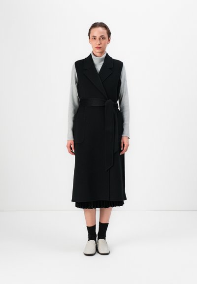 Manteau sans manches noir avec une ceinture à nouer, deux poches latérales, superposé sur un col roulé gris, assorti à des chaussettes noires et des chaussures gris clair.