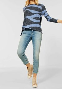 Femme portant un haut à manches longues à motifs bleu clair, un jean bleu clair court et des baskets beiges montantes, posant avec une jambe légèrement levée.