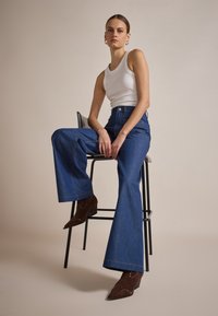 Jeans o szerokich nogawkach w niebieskim deninie z kontrastującymi szwami, w połączeniu z białym topem i brązowymi botkami ze ćwiekami, siedząc na stołku.