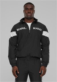 Juodas vėją atstumiančios striukės su baltais akcentais ir "KARL" logotipais ant krūtinės. Įrengta užtrauktuku, su aukštu apykakle ir elastinėmis rankovėmis.