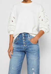 Bílá mikina s vyřezávanými detaily na rukávech, spárovaná s modrými vysokými jeansy s knoflíkovým zapínáním a předními kapsami.