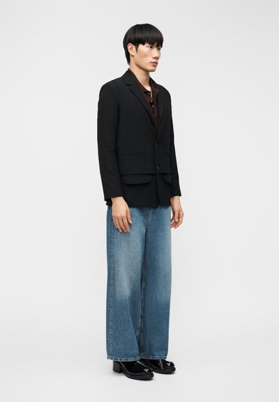 Μαύρο σακάκι με δομημένη γραμμή, σε συνδυασμό με φαρδιά τζιν παντελόνια σε ανοιχτό μπλε χρώμα. Το μοντέλο φοράει γυαλιστερά μαύρα μποτάκια στον αστράγαλο.