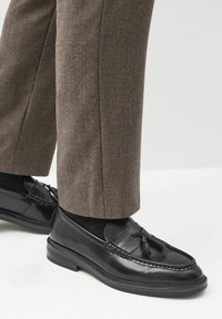 Mocassins en cuir noir avec des pampilles, des détails cousus et une semelle profilée basse, associés à un pantalon marron sur mesure.