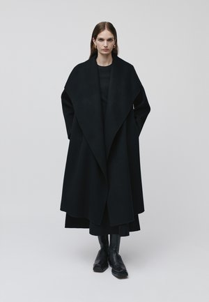 Long, black wool coat with a wide collar and draped design. Worn over a black outfit, paired with sleek black ankle boots.