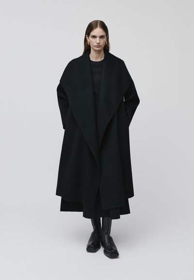 Manteau long en laine noire avec un col large et un design drapé. Porté par-dessus une tenue noire, associé à des bottines noires élégantes.