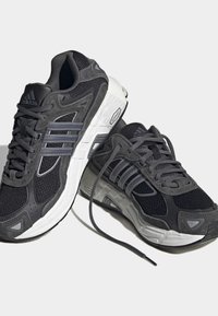 Melni un pelēki Adidas skriešanas apavi ar baltām zālēm, augšdaļa no sieta un zamša, trīs svītras sānos, viens kurpes šņorēts, otrs atstāts nesasiets.