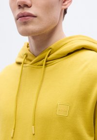 Κίτρινο φούτερ με κουκούλα φτιαγμένο από απαλό ύφασμα, με μπροστινή τσέπη, ρυθμιζόμενα κορδόνια και τετράγωνο λογότυπο "BOSS" στην αριστερή πλευρά του στήθους.