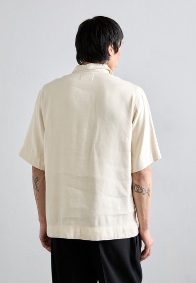 Chemise à manches courtes, couleur beige clair, avec un col, présentant un tissu texturé et un ourlet droit, vue de dos.